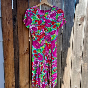 May include: A pink and green floral print dress with short sleeves and a V-neckline. The dress is hanging on a wooden hanger against a wooden fence.