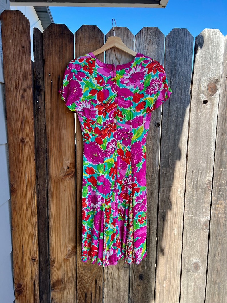 May include: A pink and green floral print dress with short sleeves and a V-neckline. The dress is hanging on a wooden hanger against a wooden fence.