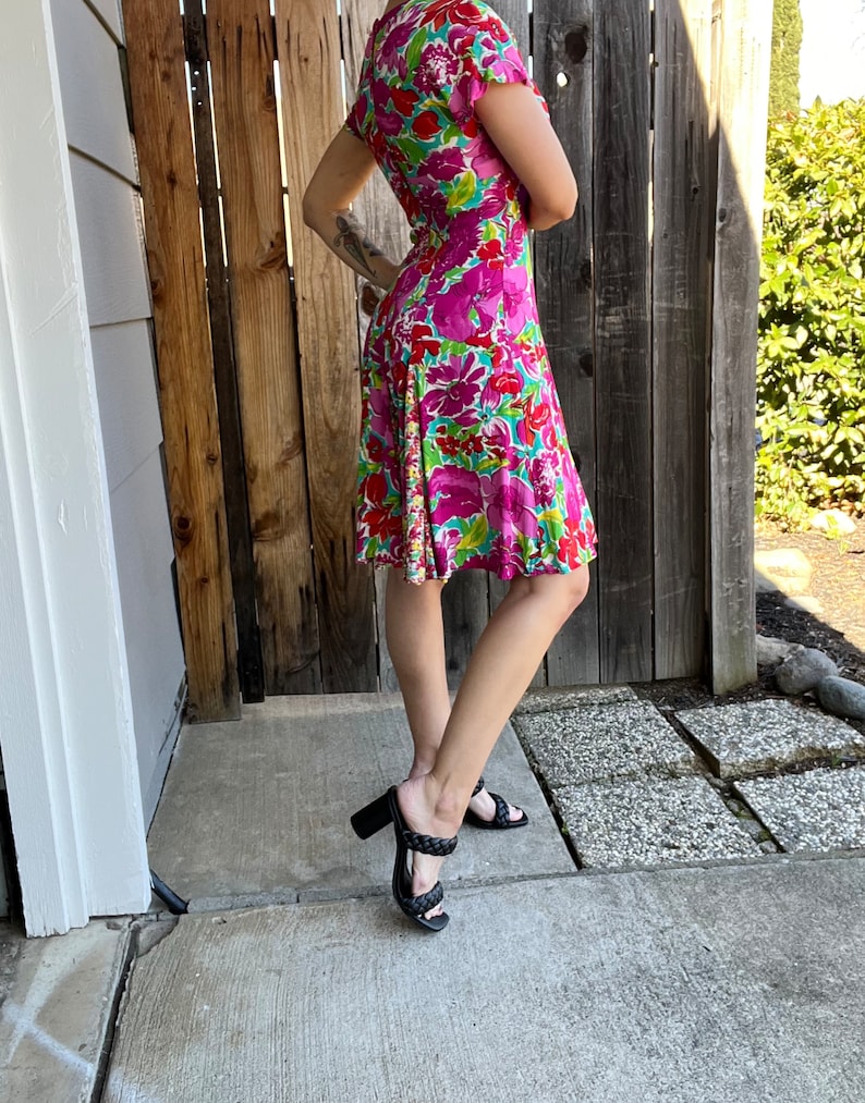 May include: A pink and green floral print dress with short sleeves and a fitted bodice. The dress has a flared skirt and is worn with black heeled sandals.