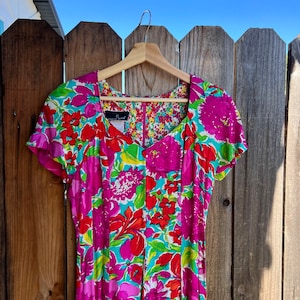 May include: A pink and green floral print dress with short sleeves and a V-neckline. The dress is hanging on a wooden hanger against a wooden fence.