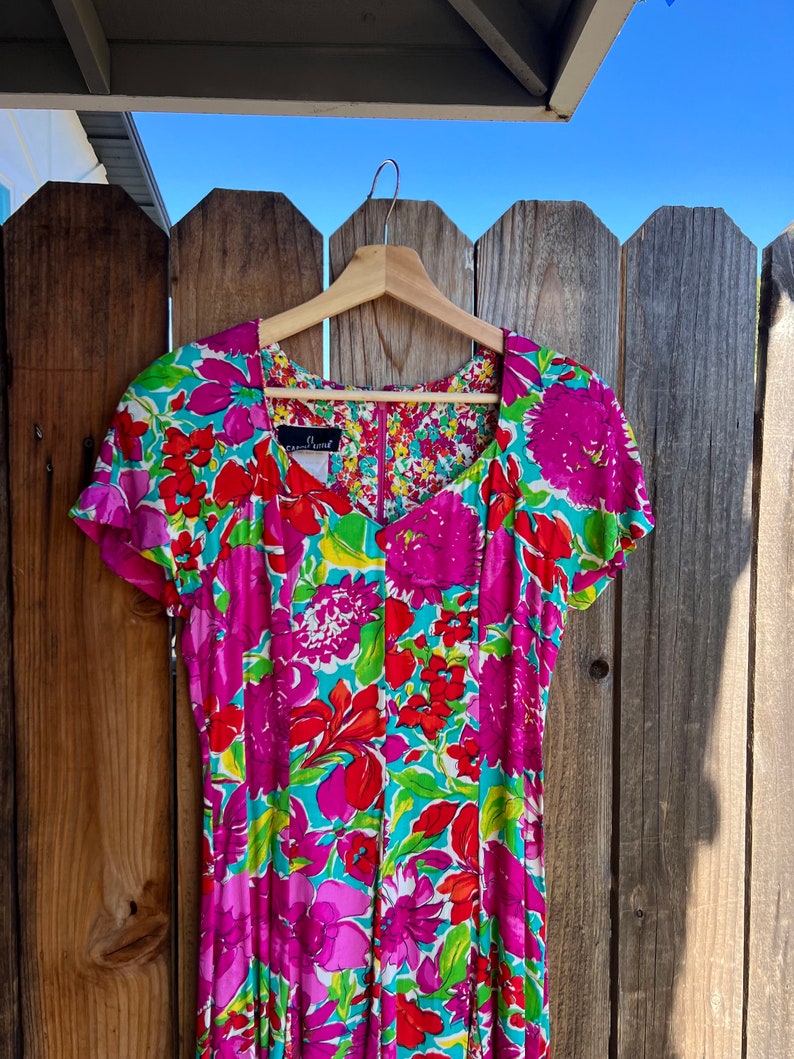 May include: A pink and green floral print dress with short sleeves and a V-neckline. The dress is hanging on a wooden hanger against a wooden fence.