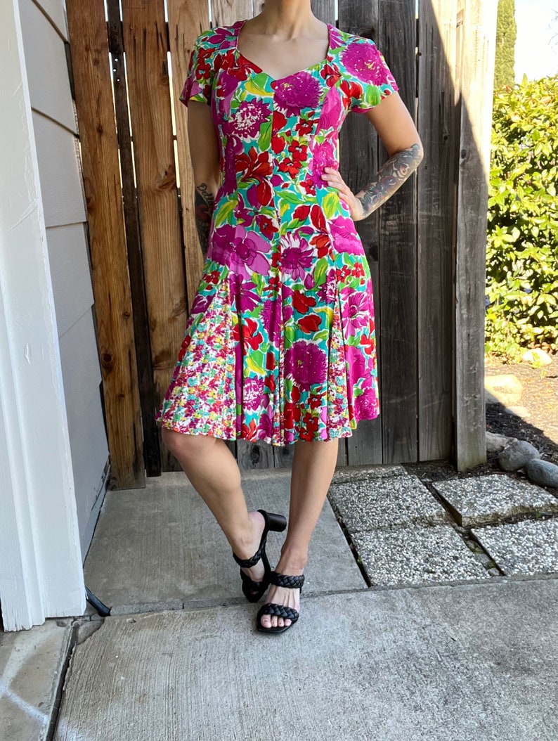 May include: A floral print dress with a pink, green, and red color scheme. The dress has a fitted bodice and a flared skirt. The dress is knee-length and has short sleeves.
