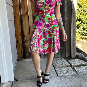 May include: A pink and green floral print dress with short sleeves and a fitted bodice. The dress has a flared skirt and a sweetheart neckline. The model is wearing black heeled sandals.