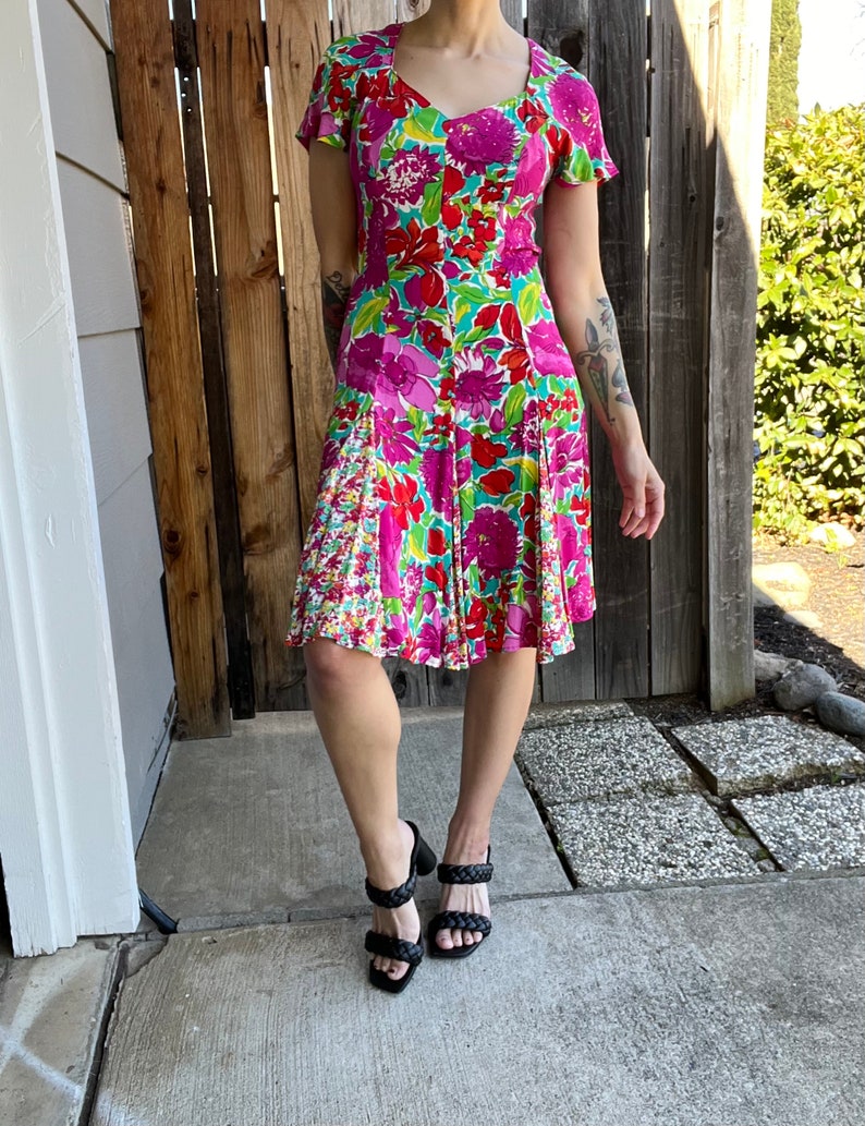 May include: A pink and green floral print dress with short sleeves and a fitted bodice. The dress has a flared skirt and a sweetheart neckline. The model is wearing black heeled sandals.