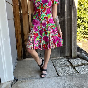 May include: A pink, green, and red floral print dress with a square neckline and short sleeves. The dress has a fitted bodice and a flared skirt.