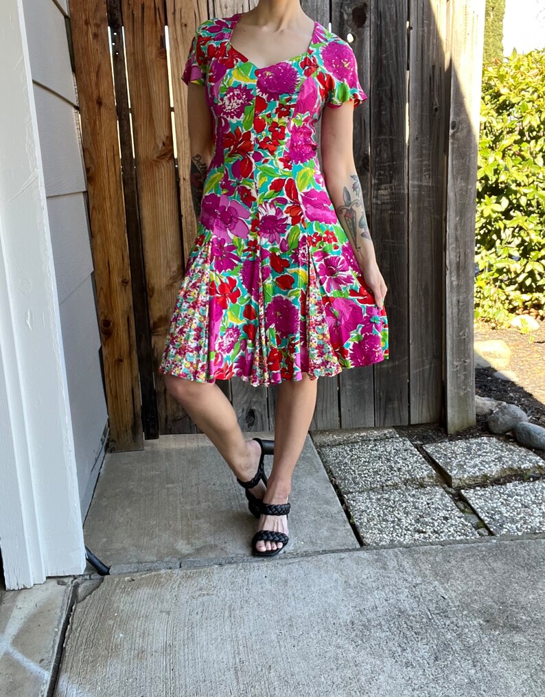 May include: A pink, green, and red floral print dress with a square neckline and short sleeves. The dress has a fitted bodice and a flared skirt.
