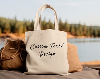 Puede incluir: Una bolsa de lona blanca con texto negro que dice "Custom Text/ Design". La bolsa está sentada sobre una superficie marrón con una bolsa de arpillera marrón detrás.