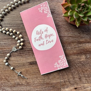 Peut inclure: Un livret rose avec le texte "Acts of Faith, Hope, and Love" est posé à côté d'un chapelet et d'une petite plante succulente en pot. Le livret a des motifs floraux blancs. Le chapelet est fait de perles claires.