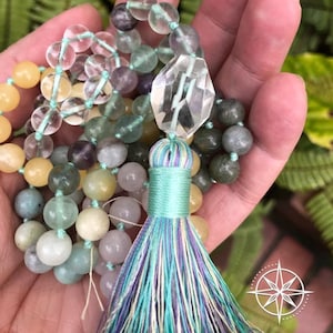 Op de afbeelding: Een veelkleurige edelsteen mala ketting met een hanger van helder kwartskristal en een turquoise kwast. De ketting is gemaakt van verschillende stenen, waaronder amethist, fluoriet en aventurijn.