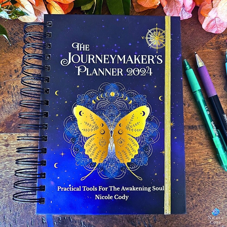 Image of the Journeymaker's Planner 2024 with  gold butterfly on a blue background. There is gold elastic, a blue page marker ribbon, spiral binding and a hard cover. It is mystical and intuitive, pretty and feminine. Magical and practical.