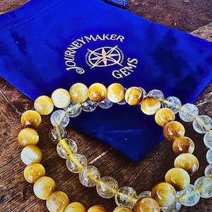 May include: Two beaded bracelets, one with golden tiger's eye beads and the other with clear quartz beads, are displayed on a blue velvet pouch with the logo "Journey Maker Gems".