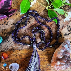 May include: A purple amethyst mala with a clear quartz bead and a silver tassel. The mala is on a wooden surface with other crystals.