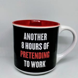 Może przedstawiać: Ceramiczny kubek z białą stroną zewnętrzną i czerwonym wnętrzem. Czarny przód zawiera biały napis "ANOTHER 8 HOURS OF", po którym następuje czerwony napis "PRETENDING" oraz biały napis "TO WORK" pogrubioną czcionką bezszeryfową.