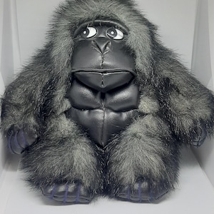 Puede incluir: Un peluche de gorila con pecho y cara de piel sintética negra, rodeado de pelo sintético largo y desgreñado en tonos gris oscuro y negro. El gorila tiene ojos grandes y expresivos en blanco y negro que miran hacia arriba y costuras moradas.