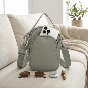 May include: A sage green crossbody bag with a white smartphone in the front pocket. The bag has a shoulder strap and silver-tone hardware. Also pictured are sunglasses, a small silver object, and a brown pen.