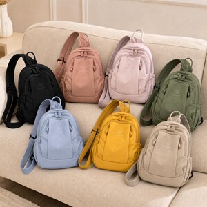 May include: A selection of small backpacks in a range of colours, such as black, pink, light blue, yellow, and green. Each bag has a single shoulder strap and a front pocket. The bags are displayed on a beige sofa.