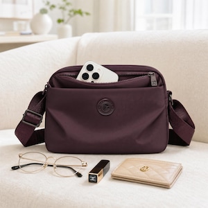 May include: A dark purple crossbody bag with a zippered main compartment containing a white smartphone. The bag features a matching adjustable fabric strap, a circular logo patch on the front, and dark metal hardware. A pair of gold-rimmed glasses, a black lipstick tube, and a beige quilted card holder are placed in front.