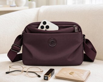 May include: A dark purple crossbody bag with a zippered main compartment containing a white smartphone. The bag features a matching adjustable fabric strap, a circular logo patch on the front, and dark metal hardware. A pair of gold-rimmed glasses, a black lipstick tube, and a beige quilted card holder are placed in front.