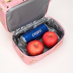 May include: Pink lunch bag with a zipper closure and a printed design of cartoon dogs. The bag is open and contains two red apples and a blue can of Pepsi.