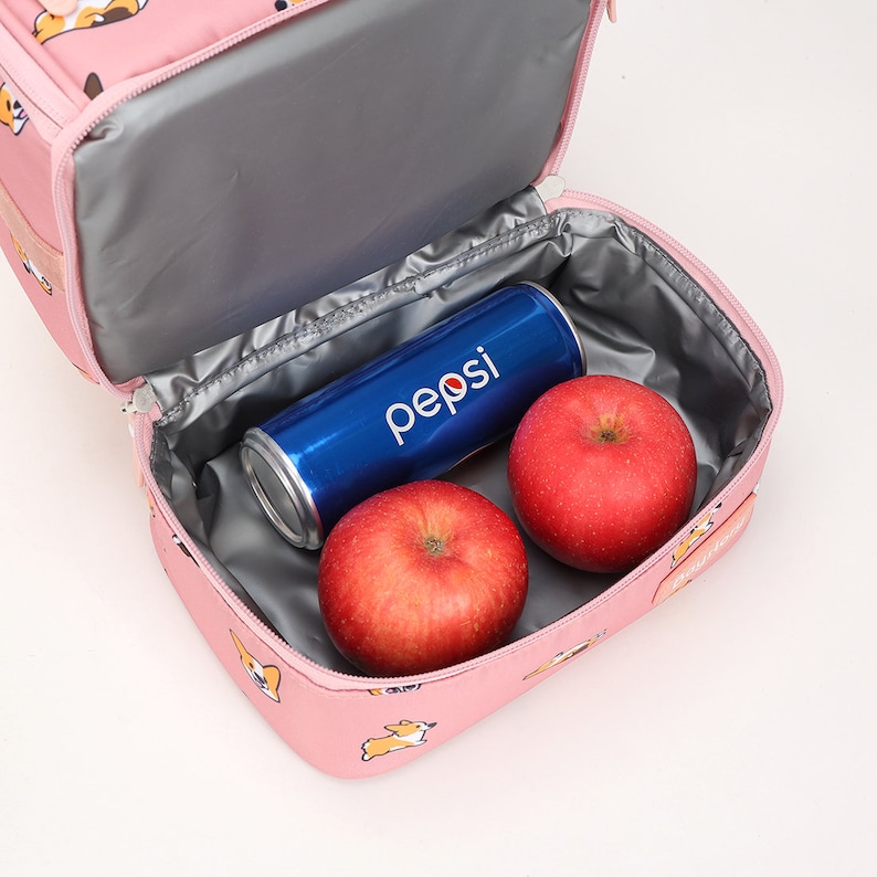 May include: Pink lunch bag with a zipper closure and a printed design of cartoon dogs. The bag is open and contains two red apples and a blue can of Pepsi.