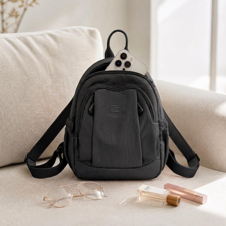 May include: A black nylon backpack with adjustable straps, a top handle, and a front pocket. A smartphone is visible inside the main compartment. The backpack is on a beige sofa, with eyeglasses, perfume, and lipstick nearby.