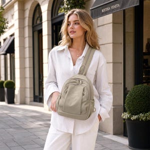 May include: A light olive green backpack with a single shoulder strap. The backpack has a rounded shape with multiple zipped compartments and a small pocket. The bag is made of a smooth, durable material.