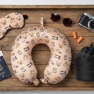 May include: A travel set on a wooden tray, including a beige neck pillow, eye mask, and a black drawstring bag, all patterned with corgi illustrations. Also included are sunglasses, a camera, earplugs, and a passport.
