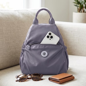 May include: A lavender backpack with a top handle, a zippered front pocket containing a white smartphone, and side elastic pockets. A circular logo patch is centered on the front. Tortoiseshell sunglasses and a brown wallet are placed in front.