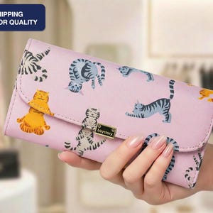 May include: A pink wallet featuring a repeating pattern of playful cat illustrations in various poses and colors. The wallet has a flap closure and a small metal clasp. The text "baystory" is visible on the clasp.