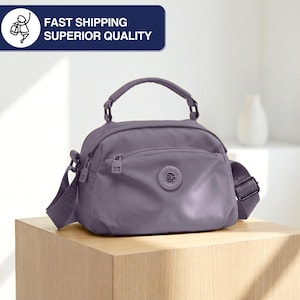 May include: A lavender-colored crossbody bag with a top handle and adjustable shoulder strap. The bag has a front zippered pocket and a circular emblem. The text "FAST SHIPPING SUPERIOR QUALITY" is in a blue rectangle.