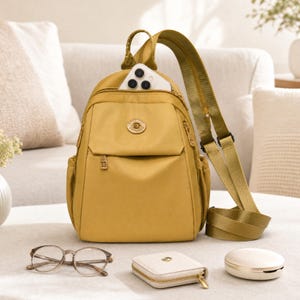 May include: A mustard yellow nylon backpack with a top handle and adjustable shoulder straps. The front features a zippered pocket with a circular gold logo emblem. A white smartphone is tucked into the top compartment, and the bag has side pockets.