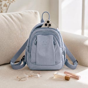 May include: A pale blue backpack with a top handle and adjustable straps. The backpack has a front pocket and a main compartment with a smartphone inside. A pair of gold-rimmed glasses, perfume, and a lipstick are next to the backpack.