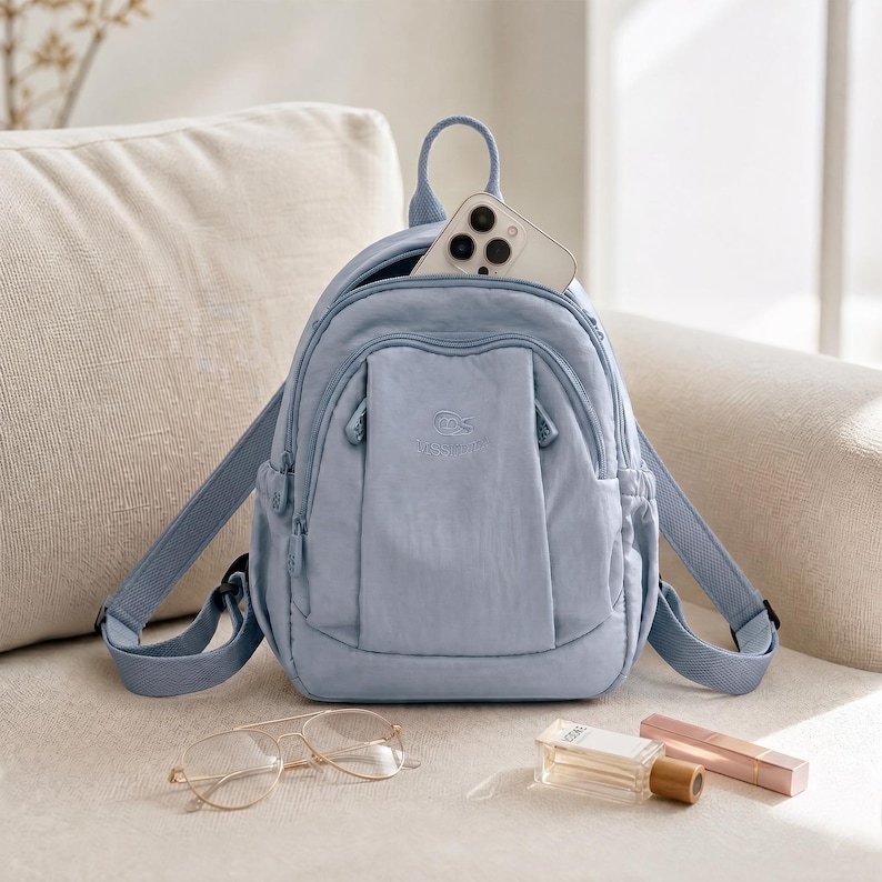 May include: A pale blue backpack with a top handle and adjustable straps. The backpack has a front pocket and a main compartment with a smartphone inside. A pair of gold-rimmed glasses, perfume, and a lipstick are next to the backpack.