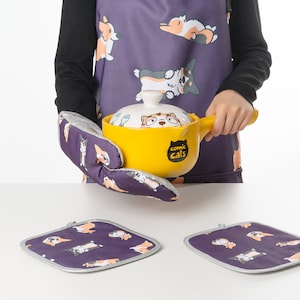 May include: A purple oven mitt with a cartoon cat design and the text "comic cats" is holding a yellow ceramic pot with a cat design. Two purple pot holders with a cartoon cat design are on a white surface.