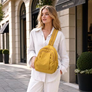 May include: Mustard yellow sling backpack with a single shoulder strap. The bag has multiple zippered compartments and a curved design. The person is wearing a white button-down shirt and white trousers. The background includes a building with arched windows.
