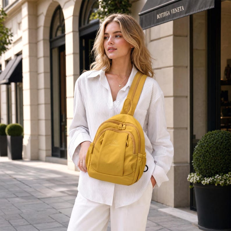 May include: Mustard yellow sling backpack with a single shoulder strap. The bag has multiple zippered compartments and a curved design. The person is wearing a white button-down shirt and white trousers. The background includes a building with arched windows.