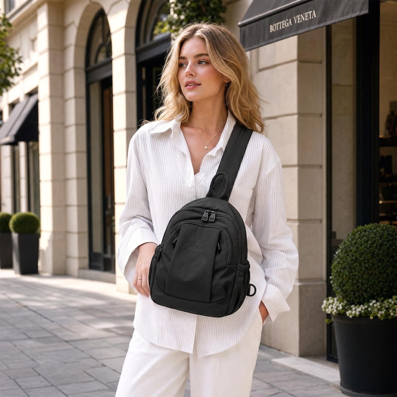 May include: A black sling backpack with a curved design, featuring a single shoulder strap and multiple zipped compartments. The bag is made of a durable material and is worn across the body. The background includes a building facade and greenery.