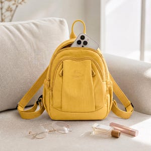 May include: A mustard yellow backpack with a top handle and adjustable straps. A smartphone is visible in the open top pocket. The backpack is on a cream sofa, with glasses, perfume, and lipstick nearby.