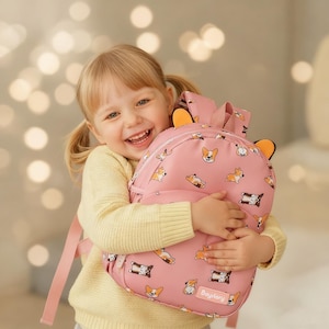 May include: Pink backpack with a repeating pattern of corgi dogs in various poses. The backpack has orange ear-shaped accents and the brand name "Baytory" printed on the bottom. The backpack is being held by a smiling child.