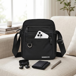 May include: Black crossbody bag with multiple zippered compartments, a silver D-ring, and a long adjustable strap. A white smartphone is in the front pocket. The bag is on a couch with a wallet, pen, and wireless earbuds.