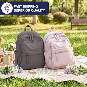 May include: Two backpacks, one gray and one pink, are displayed outdoors on a blanket. Each backpack has multiple zippered compartments and a top handle. The image also includes a picnic basket, flowers, and the text "FAST SHIPPING SUPERIOR QUALITY."