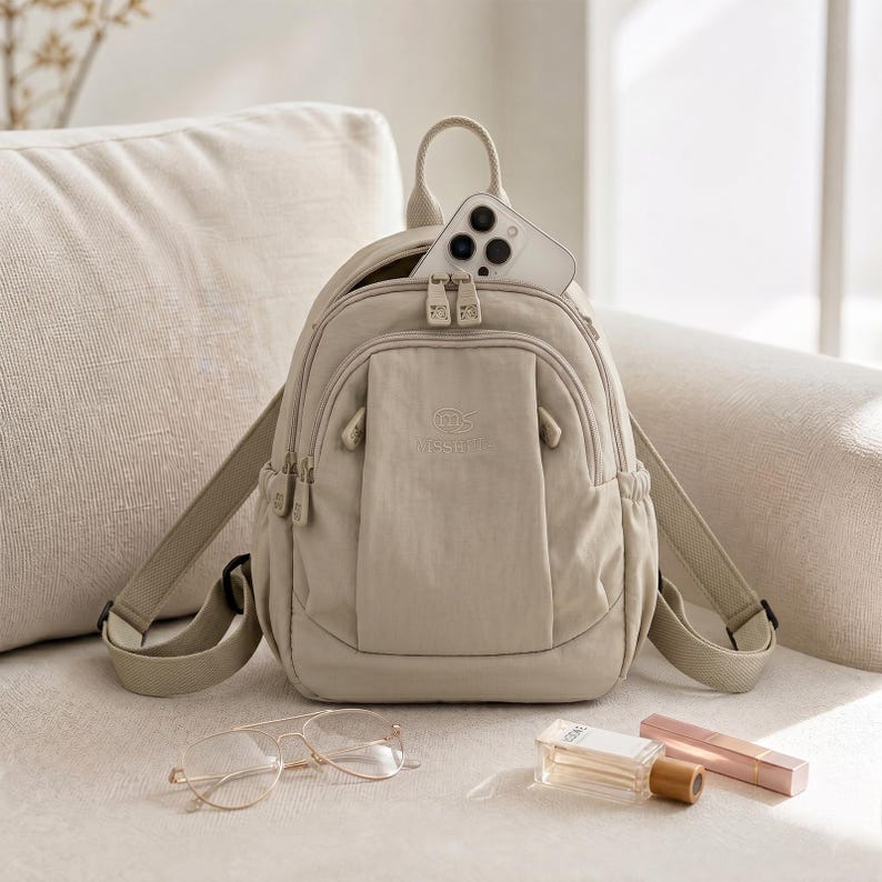 May include: A beige backpack with a top handle and adjustable straps. A smartphone is in the main compartment. Gold-rimmed glasses, a perfume bottle, and a lipstick are next to the backpack. The backpack has a small front pocket.