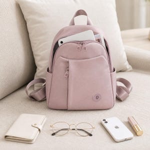 May include: A dusty rose backpack with a top handle, two shoulder straps, and a front zippered pocket. The main compartment is open, revealing a white tablet inside. A small circular logo is on the lower right side of the front pocket.
