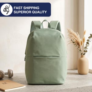 May include: A sage green backpack with a minimalist design. The backpack has a front zippered pocket and padded shoulder straps. The top of the bag has a handle for carrying. The text "FAST SHIPPING SUPERIOR QUALITY" is displayed on a blue rectangle.