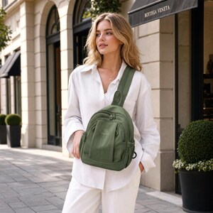 May include: An olive green sling backpack with a curved design, featuring a zippered main compartment and a smaller front pocket. The bag has a single adjustable strap and a small black loop on the side. The background includes a building facade and greenery.
