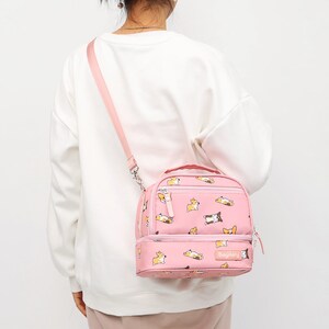 May include: Pink lunch bag with a pattern of cartoon corgis. The bag has a top handle and a shoulder strap. The bag has two compartments and a zipper closure. The bag has the word "Baylery" printed on the front.