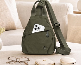 May include: An olive green sling backpack with a single adjustable shoulder strap, a top handle, and a front zipped pocket containing a white smartphone. The bag features a small circular logo patch on the front and a smooth, durable fabric exterior.