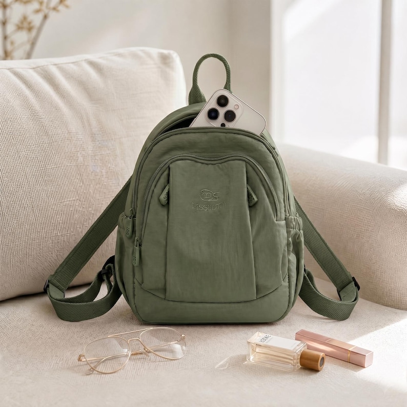 May include: An olive green backpack with adjustable straps and a top handle. A smartphone is visible in the open top pocket. The backpack is on a beige sofa, with eyeglasses, perfume, and a lipstick nearby.