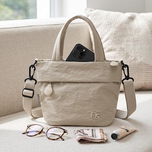 May include: A beige nylon tote bag with two top handles and a detachable shoulder strap. The bag features a black smartphone inside, a small logo on the front, and a zipper pull. Nearby are tortoise-shell glasses, a folded patterned scarf, and a tube.