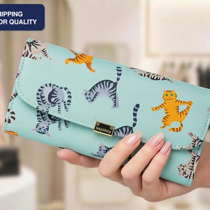May include: A light blue wallet with a cat pattern. The wallet features various cat illustrations in orange, gray, and black. The wallet has a flap closure and a gold-colored clasp. The text "FAST SHIPPING SUPERIOR QUALITY" is in the upper left corner.